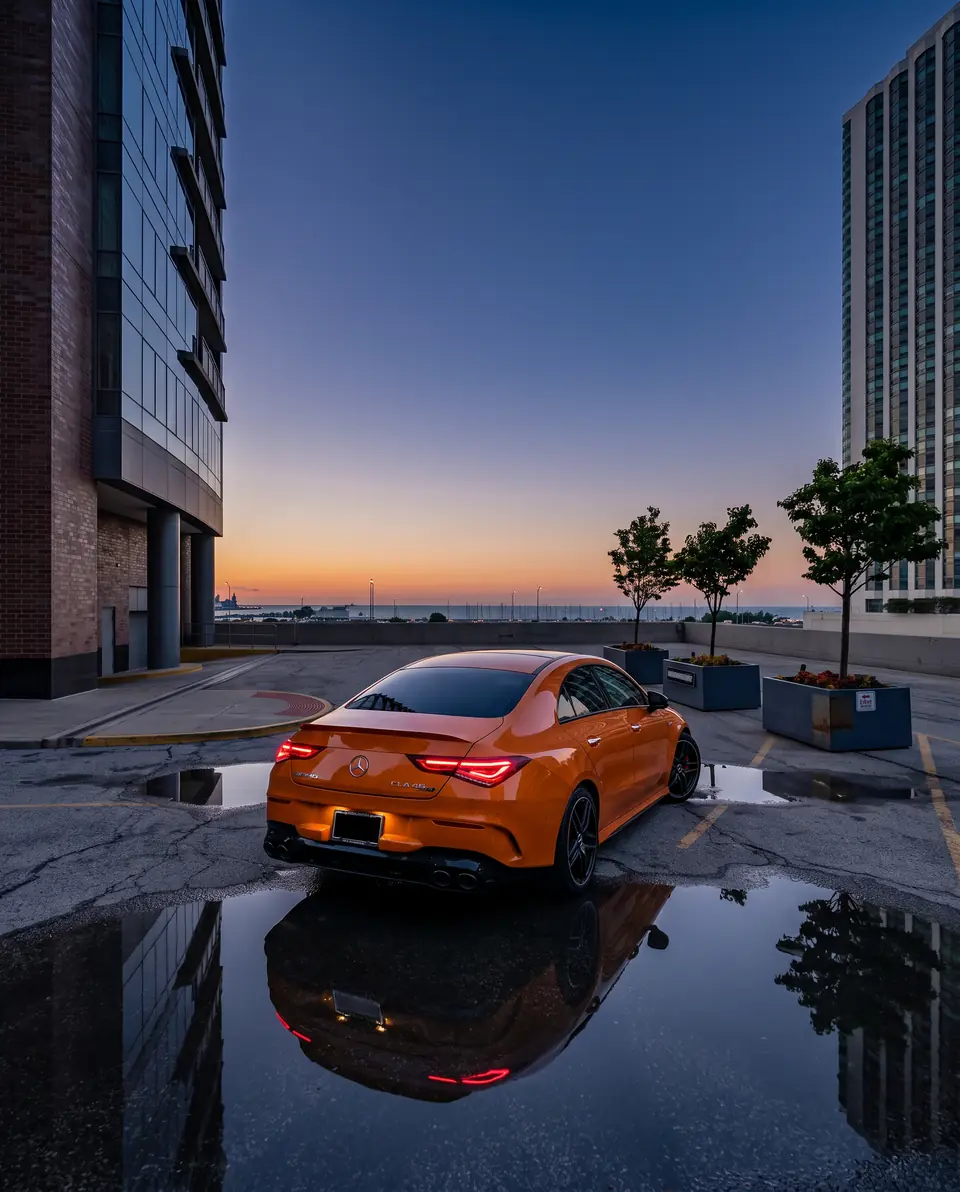An orange coupe reflected in a rain puddle on a rooftop car park at sunset